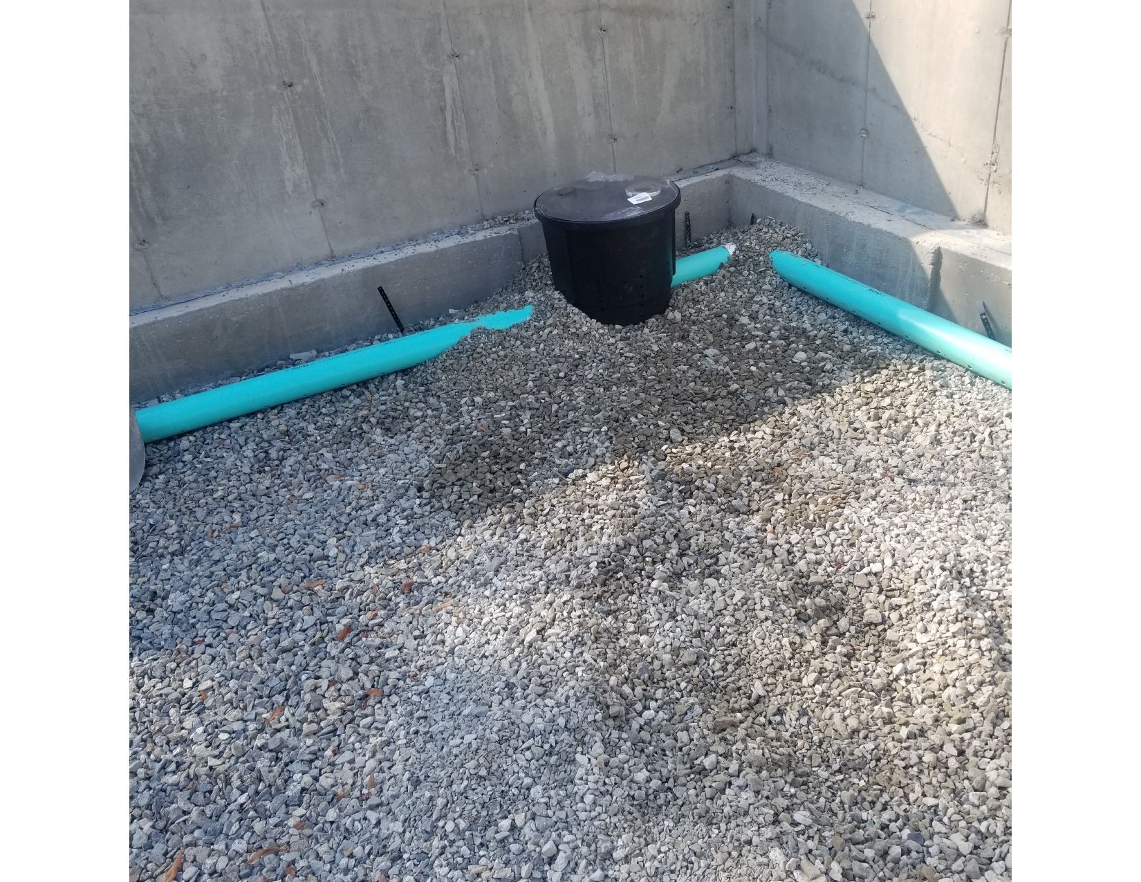 Black drainage basin, two blue pipes, and gravel in a concrete-walled foundation.