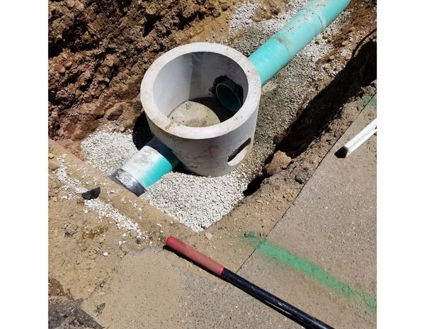 Concrete manhole and pipes in a construction site trench, surrounded by gravel and dirt.