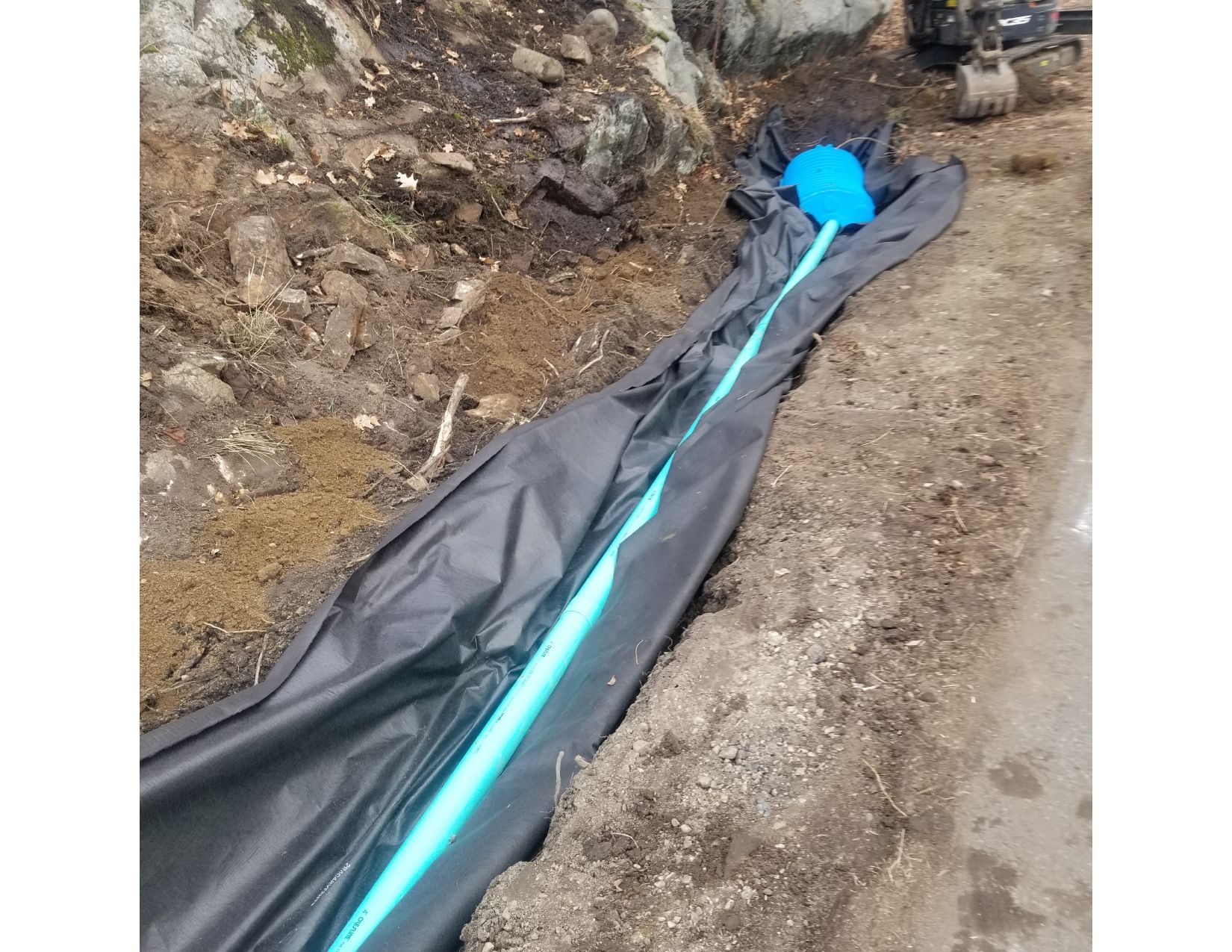 Black fabric-lined trench with a blue pipe, next to a rocky slope and a road.