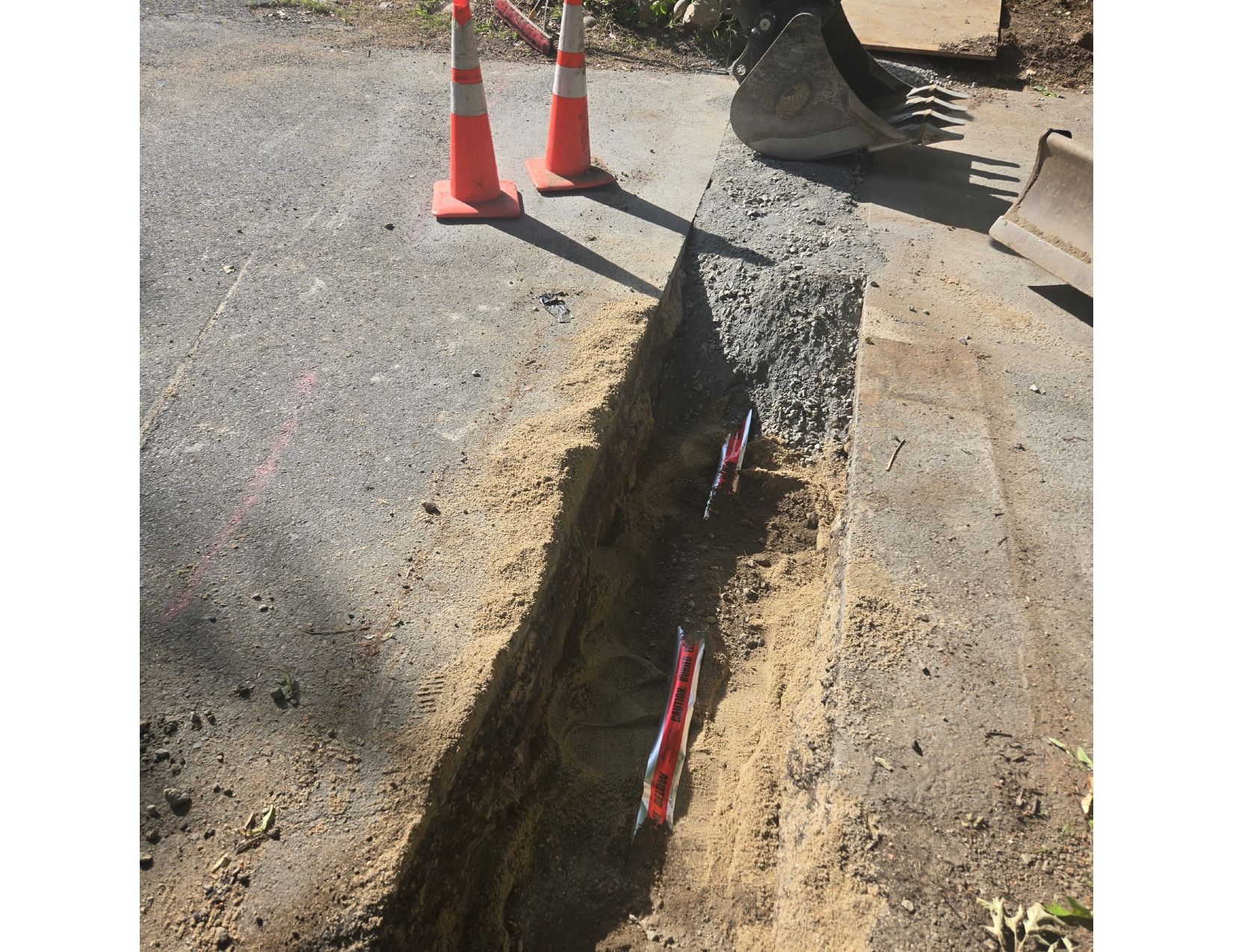 Trench dug in asphalt with orange cones. A backhoe is in the background.