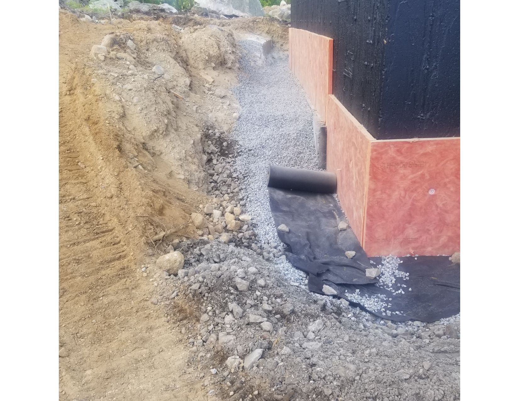 Construction site: soil mound beside a building foundation with black waterproof membrane and white gravel.