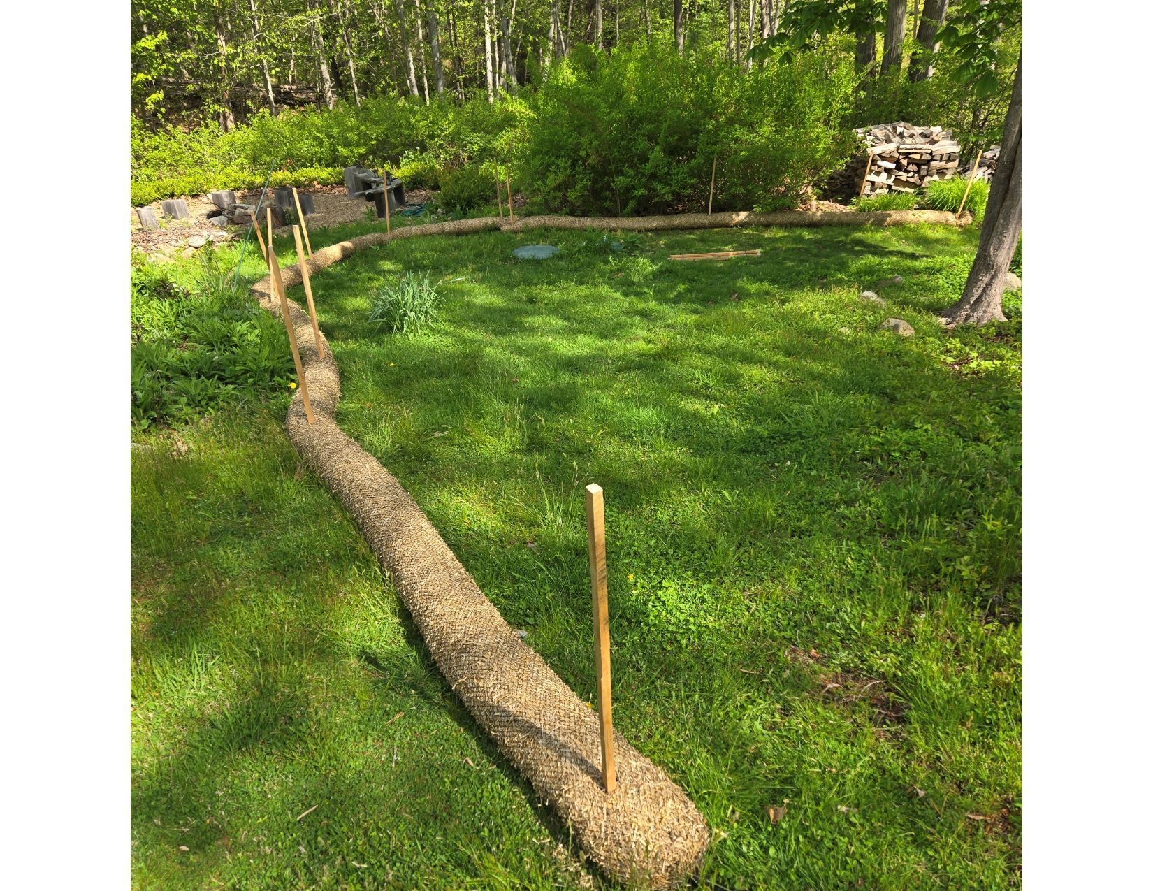 Erosion control barrier of straw logs and stakes on grassy lawn, in a shaded outdoor setting with trees and foliage.