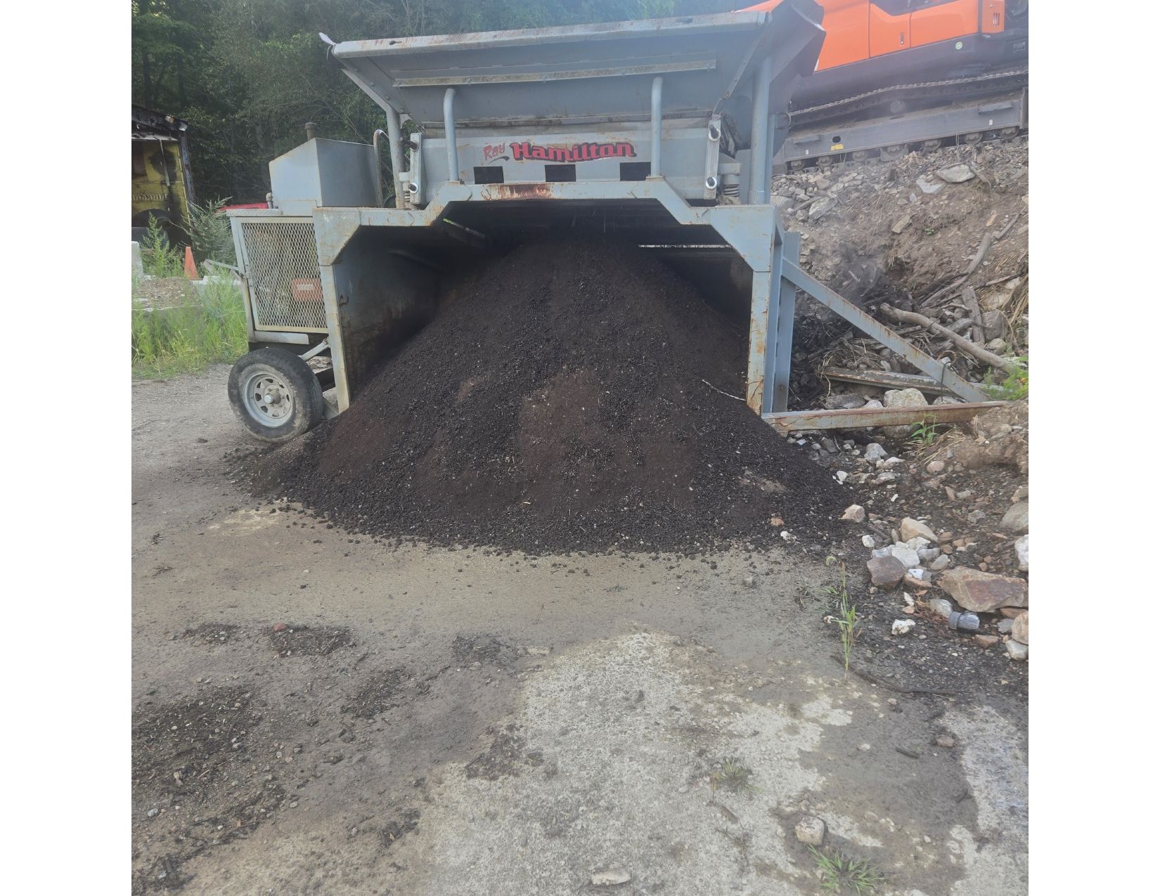 Gray machine dispensing a large pile of dark material onto a paved area; possible soil or aggregate.