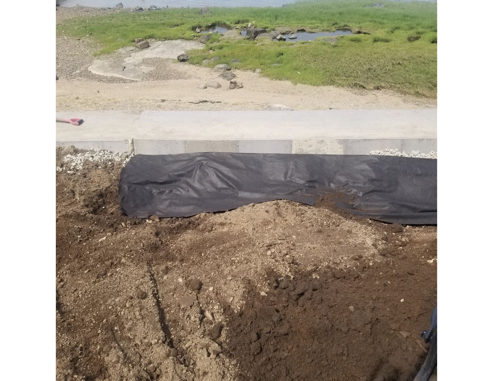 Black fabric and dirt cover an area next to a paved path and grass, with a background of water and land.