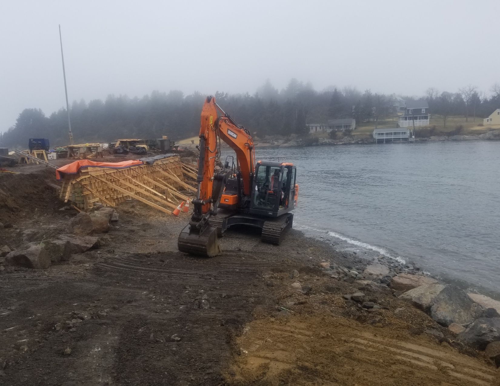Orange excavator on muddy shoreline by water with construction in background on a foggy day.