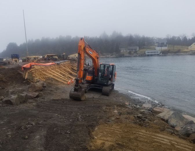 Orange excavator on muddy shoreline by water with construction in background on a foggy day.