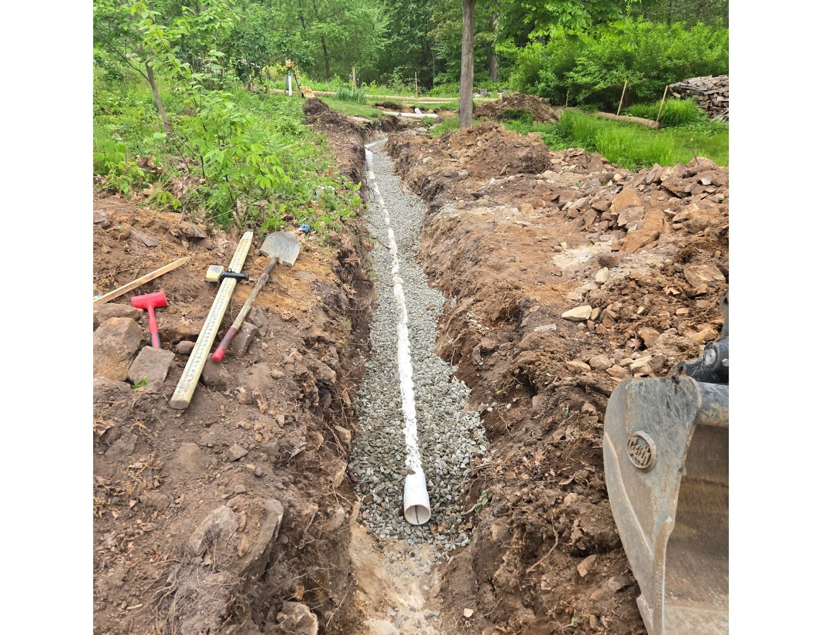 A trench dug for drainage, containing a white pipe and gravel. Soil and tools lie nearby.