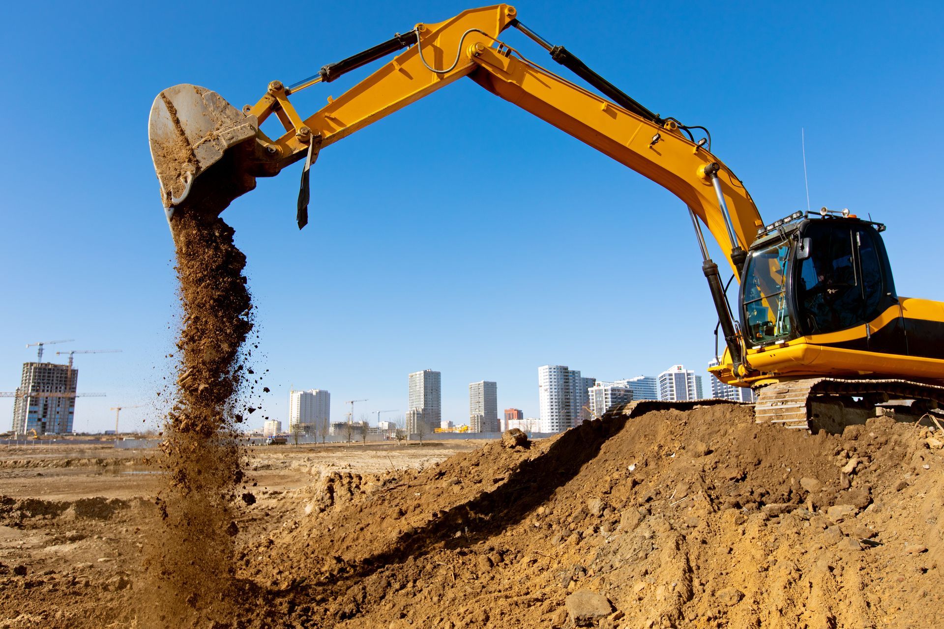 Yellow excavator dumping dirt on a construction site with city buildings in the background.