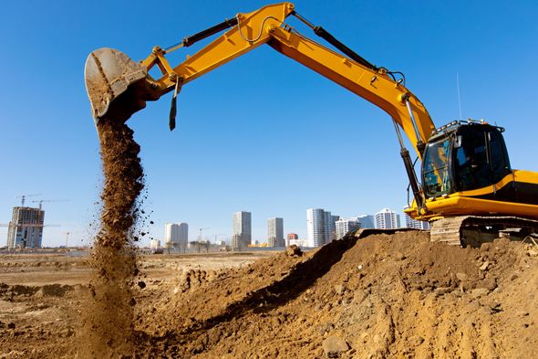 Yellow excavator dumping dirt on a construction site with city buildings in the background.