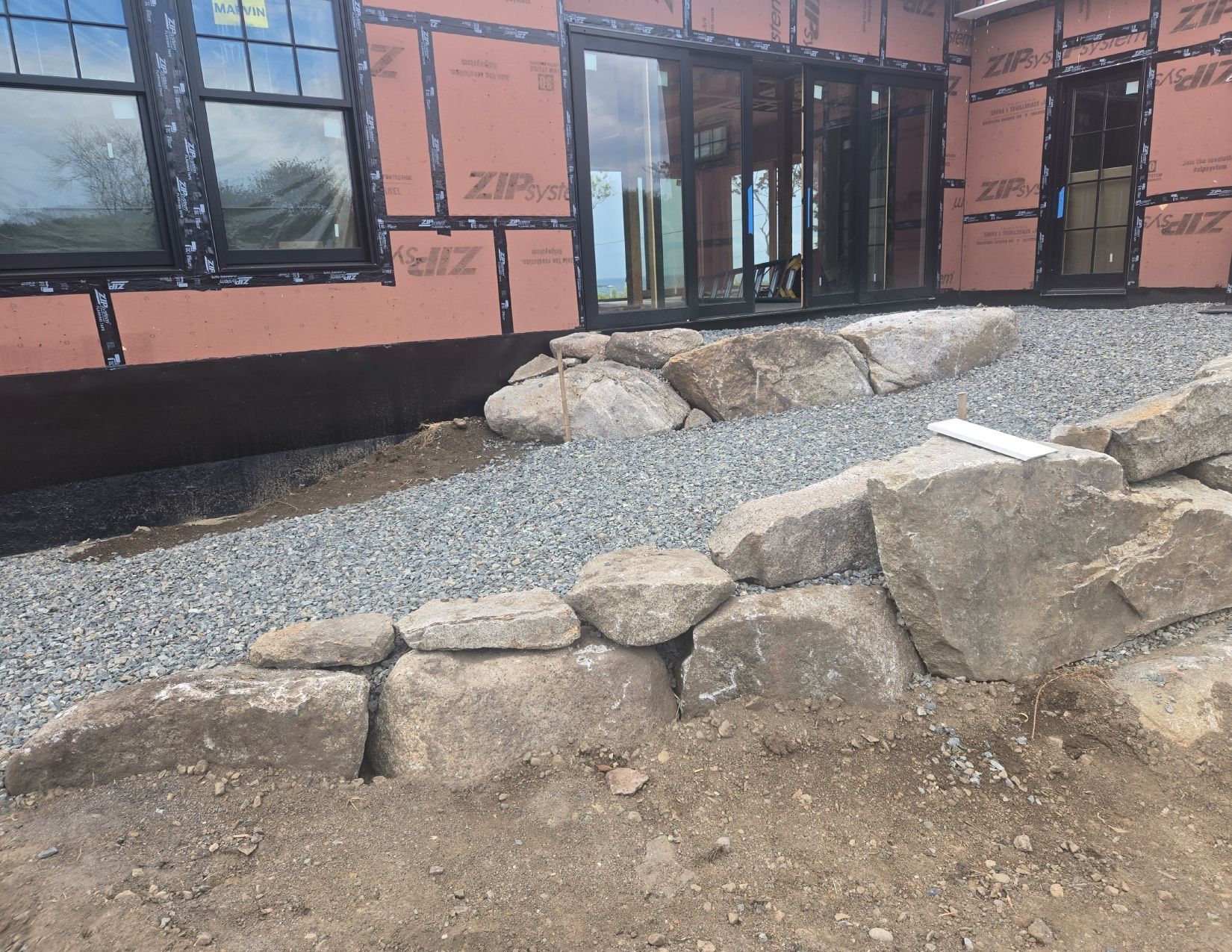 Exterior of a house under construction; large stones create a retaining wall bordering a gravel pathway.