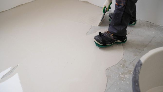 Worker applying smooth gray flooring with a trowel near a bucket and boots