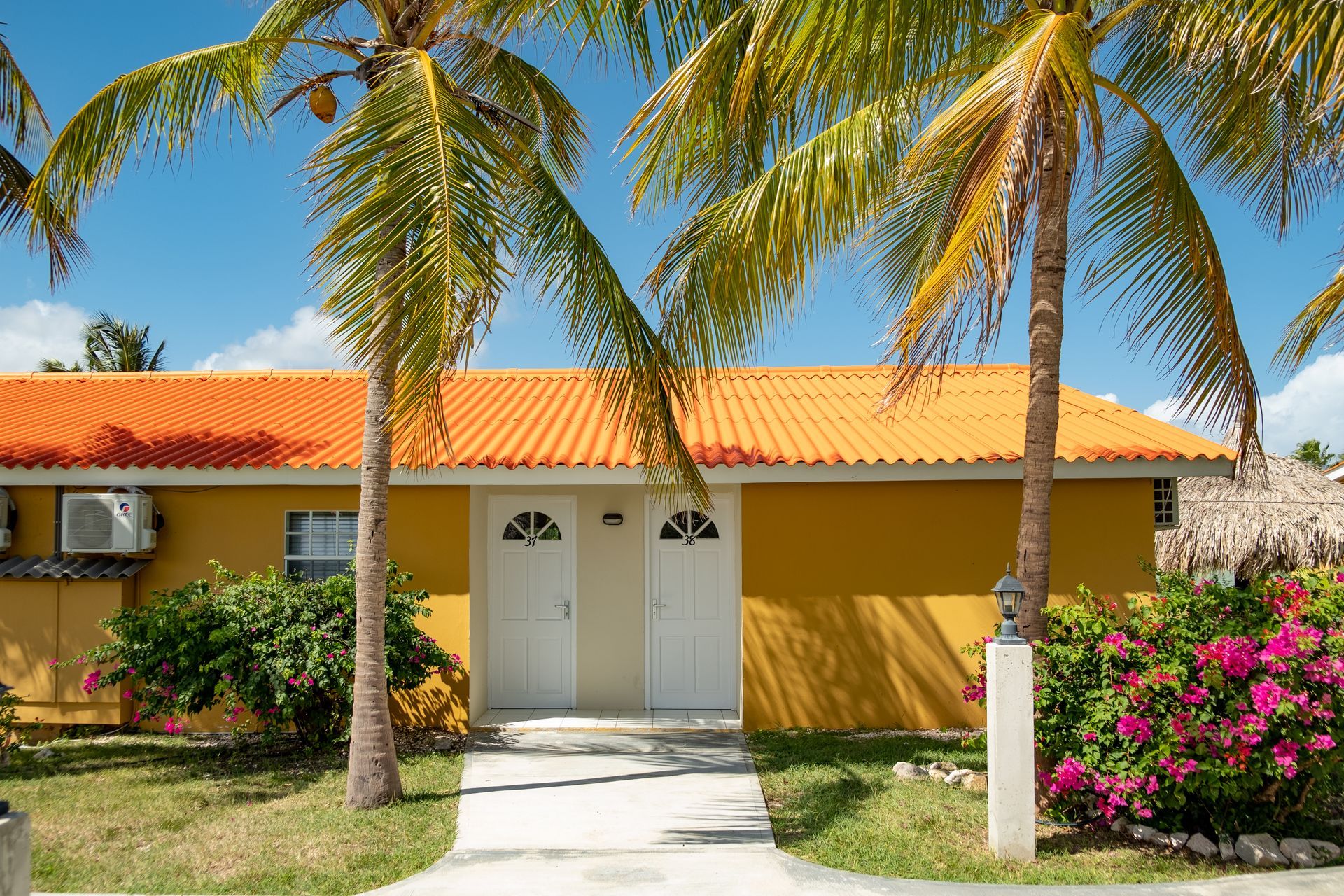 Yellow building with orange roof, white doors, palm trees, and pink flowers under a blue sky.