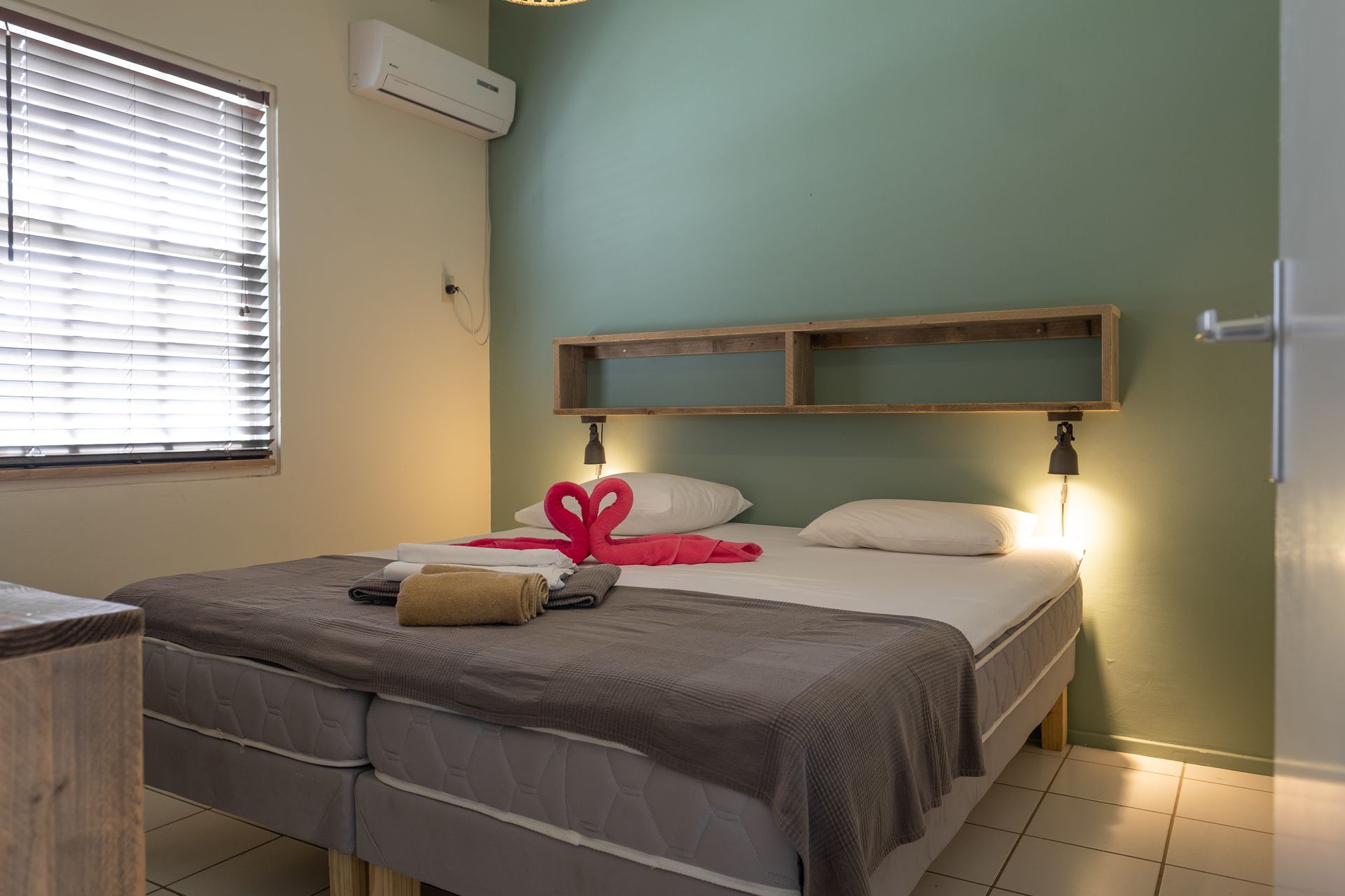 Bedroom with double bed, teal accent wall, wooden shelf, and window with blinds.
