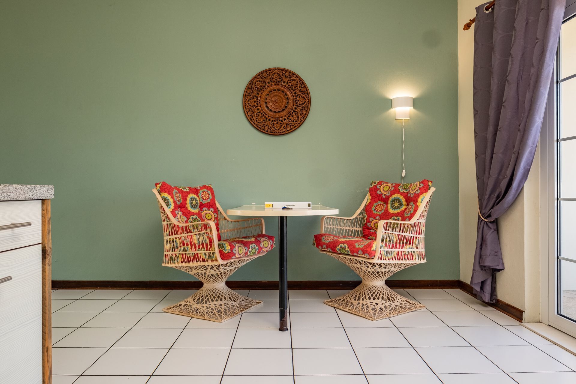 Two red patterned chairs face a small table against a green wall with a decorative circle and a lamp.
