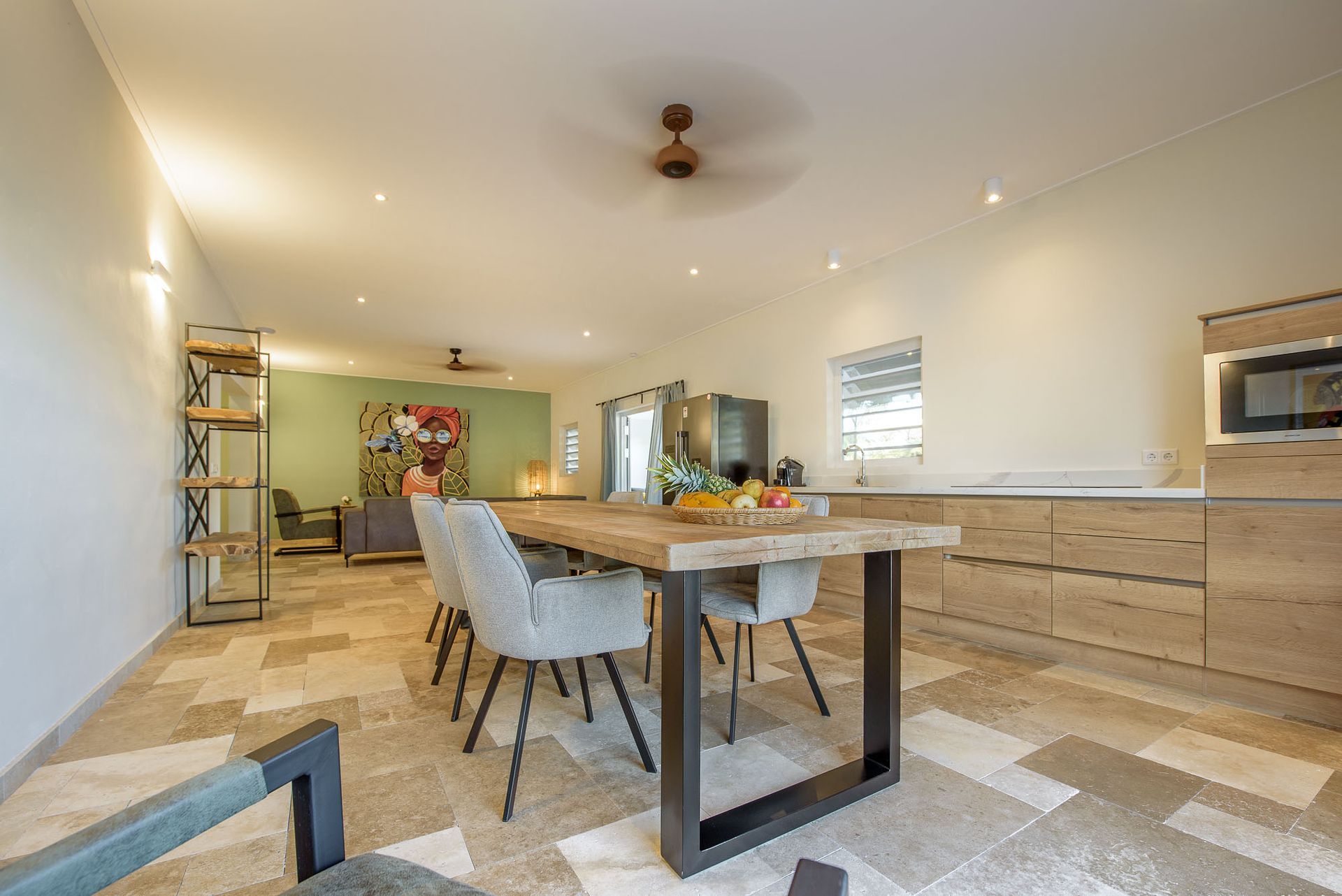 Spacious, modern dining room and kitchen with wooden table, chairs, and light-colored cabinetry; stone floors.