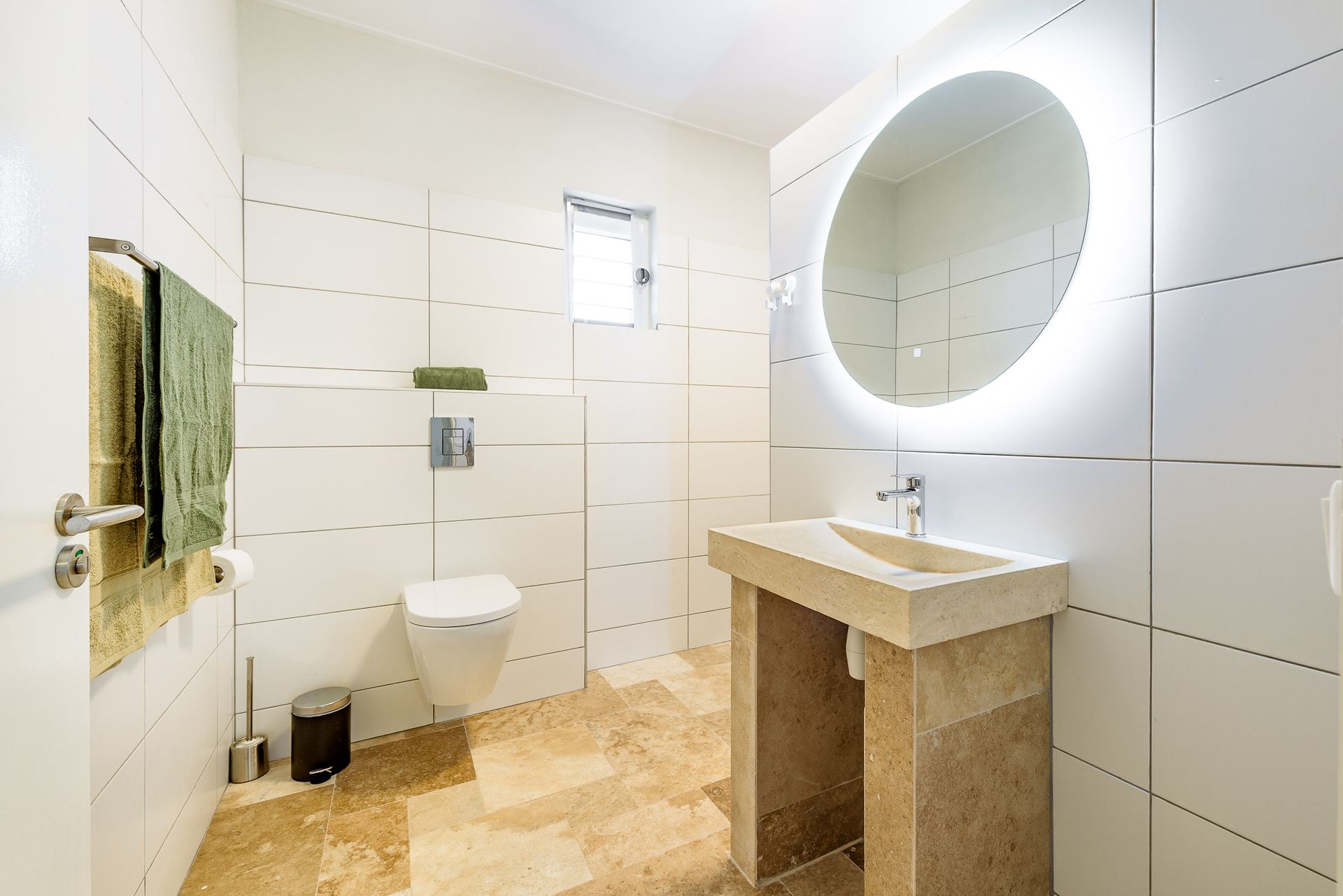 Modern white bathroom with a stone sink, round mirror with lights, and a wall-mounted toilet.