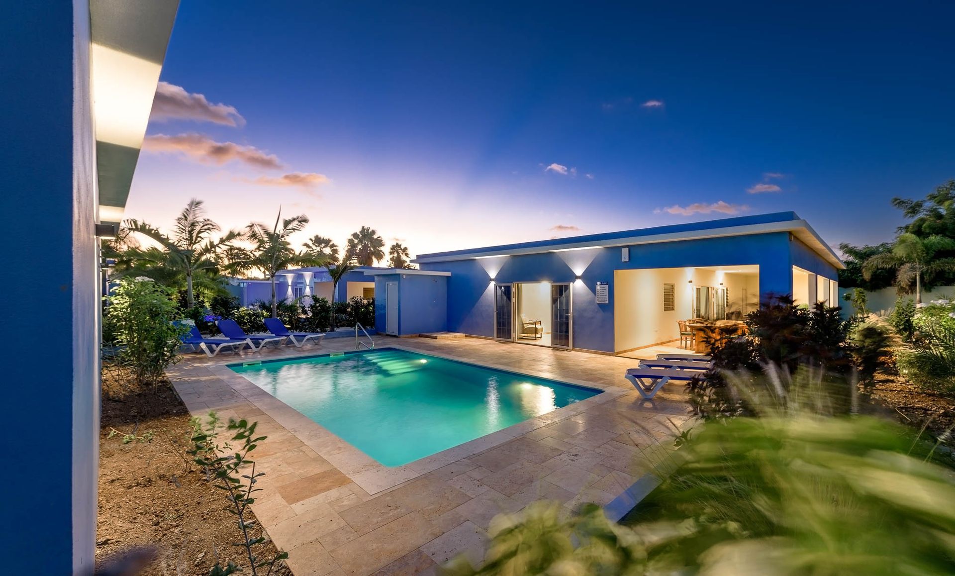 Blue house with a pool at dusk, lounge chairs by the water.