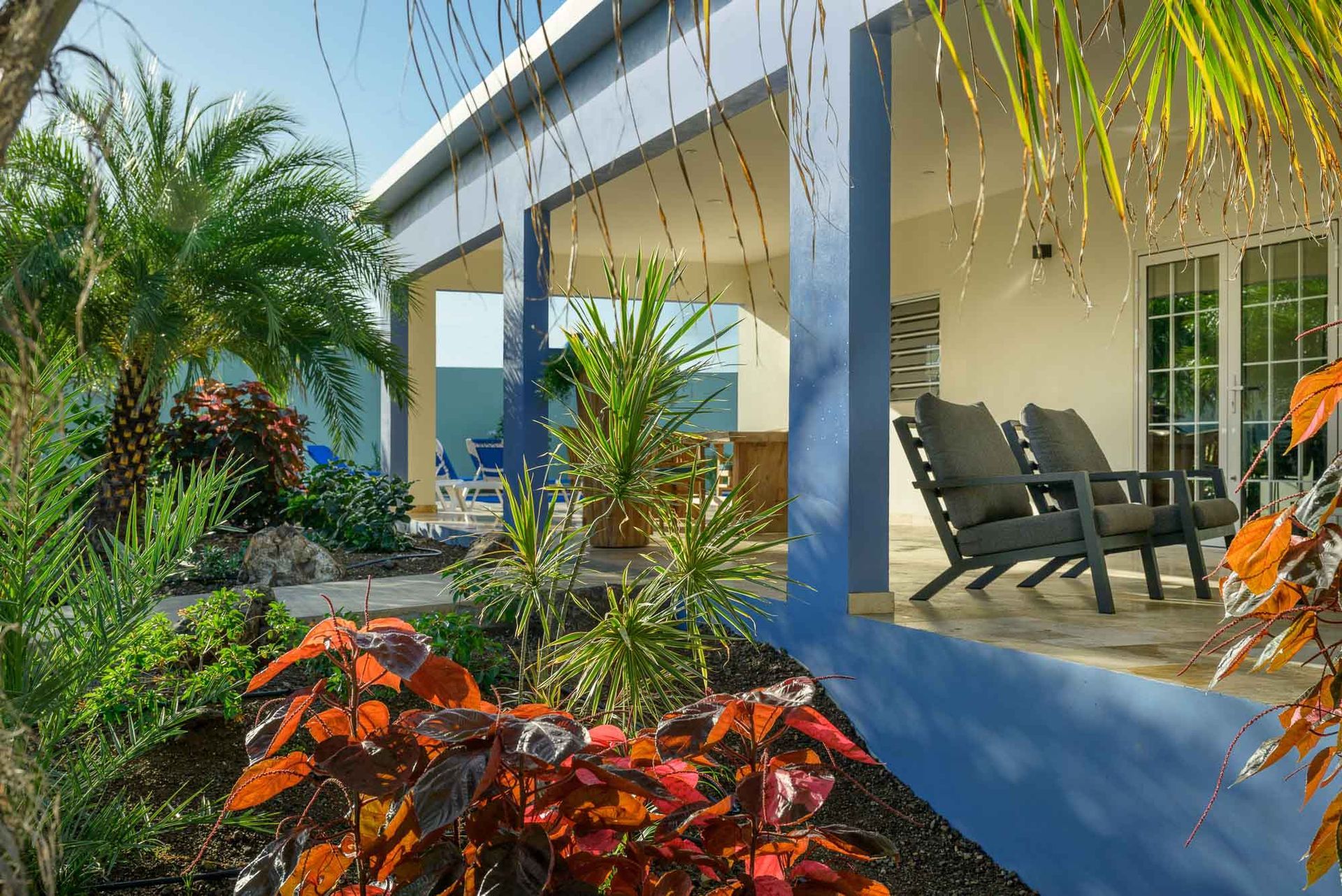 Blue house with porch, garden, and two dark chairs. Bright red and green plants are in the foreground.