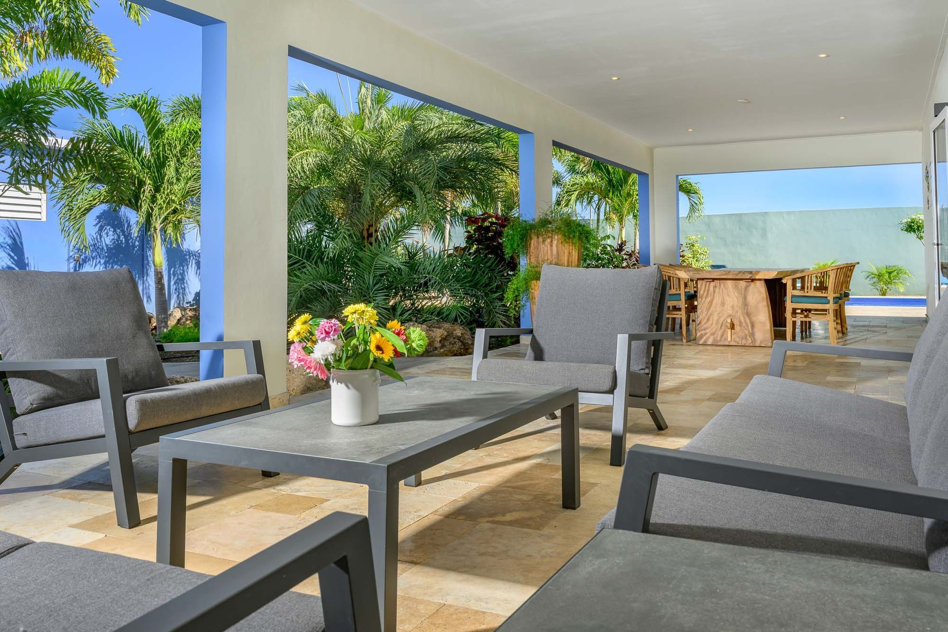 Covered patio with gray outdoor furniture and colorful flowers, overlooking a garden.