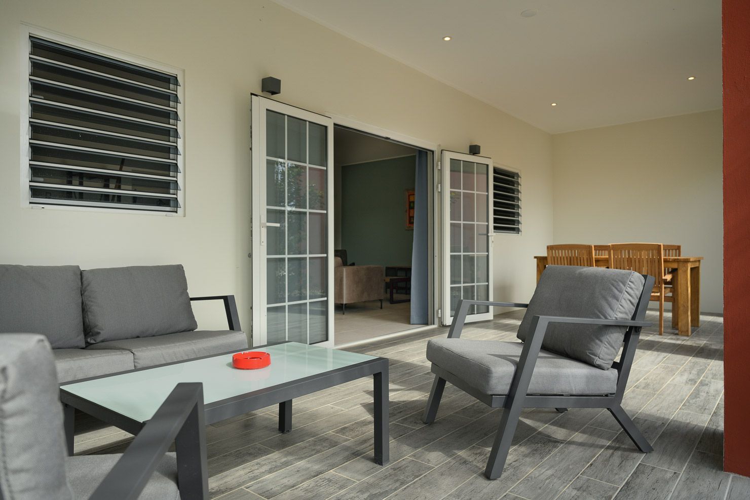 Outdoor patio with gray furniture, a glass table, and a dining table; doors lead inside.