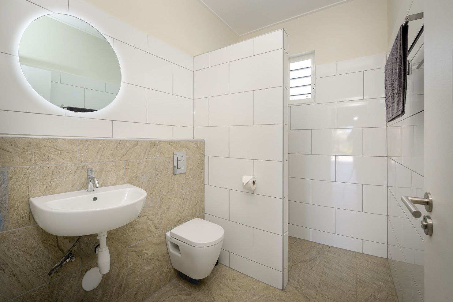 Modern bathroom with a round mirror, white tiles, and a wall-mounted sink and toilet.