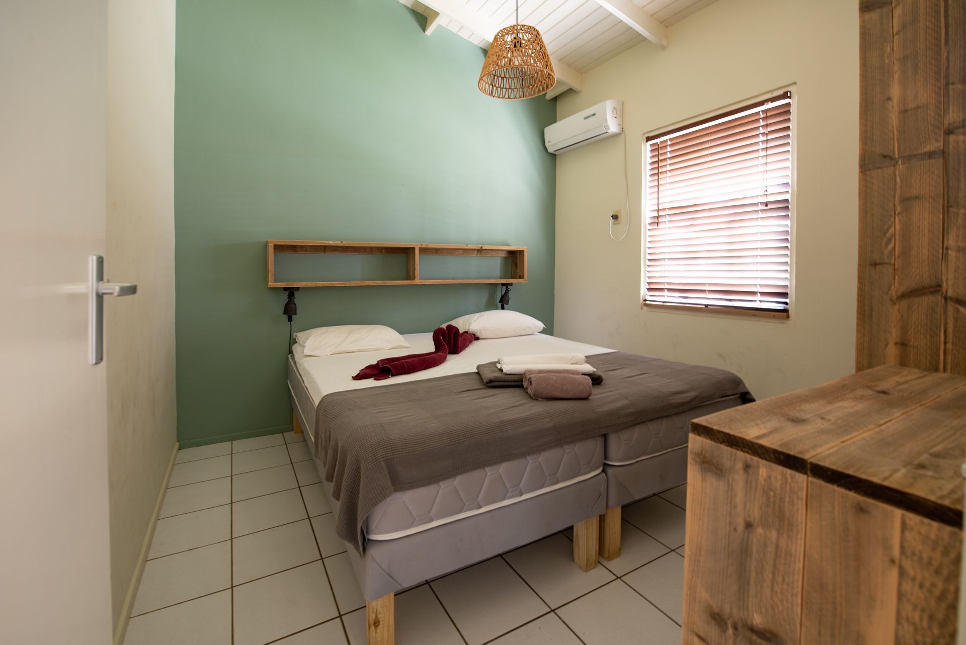 Bedroom with double bed, green wall, wooden furniture, and window with blinds.