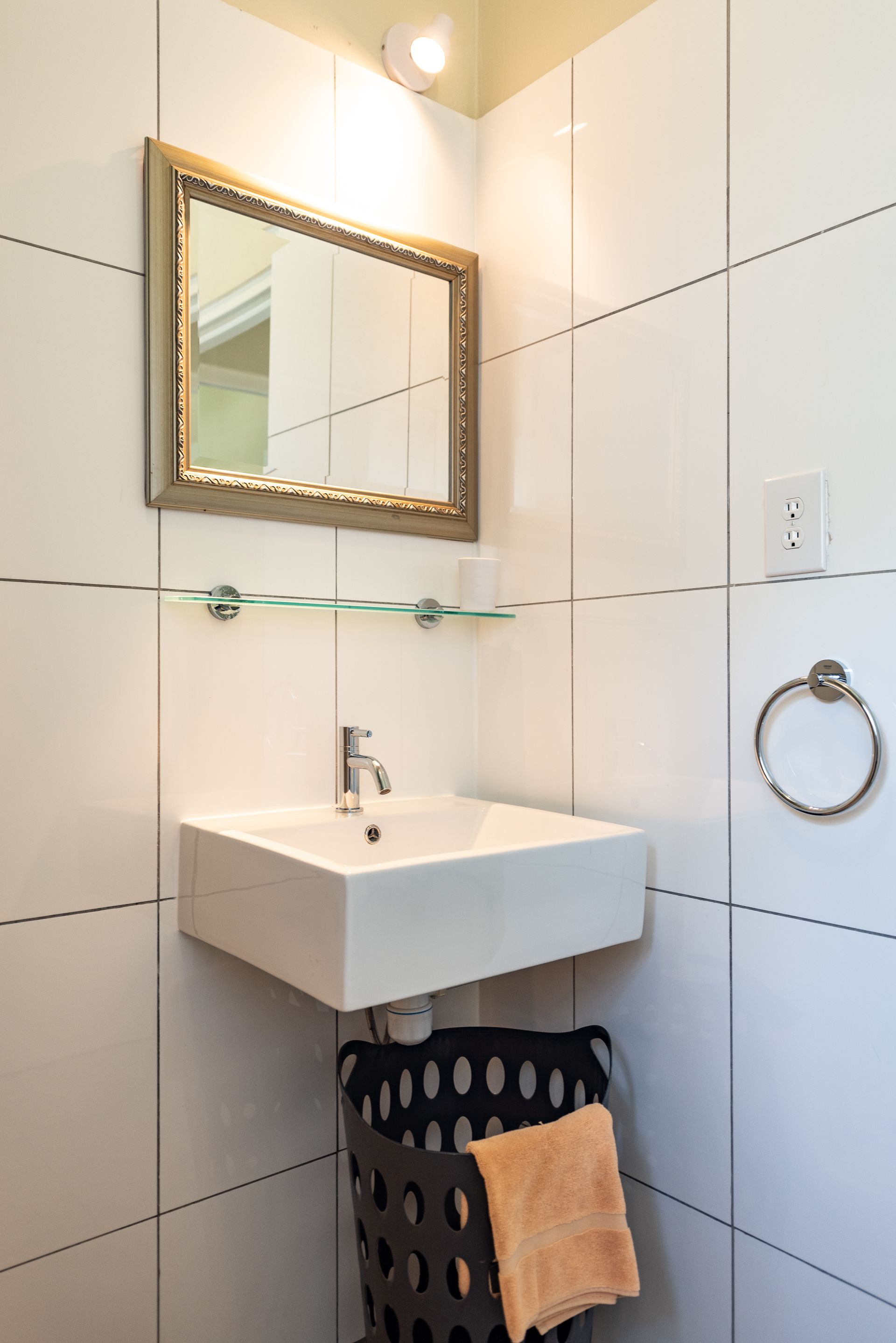 Small bathroom with white tiled walls, a square sink, mirror, and black basket with towel.