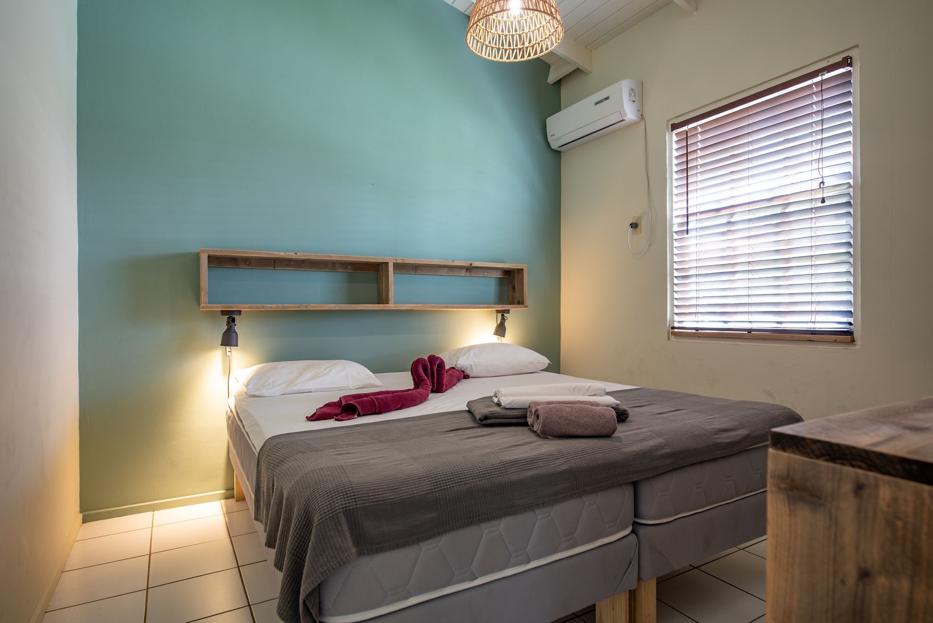 Bedroom with two beds, green and beige walls, wooden shelves, air conditioning, and a window with blinds.
