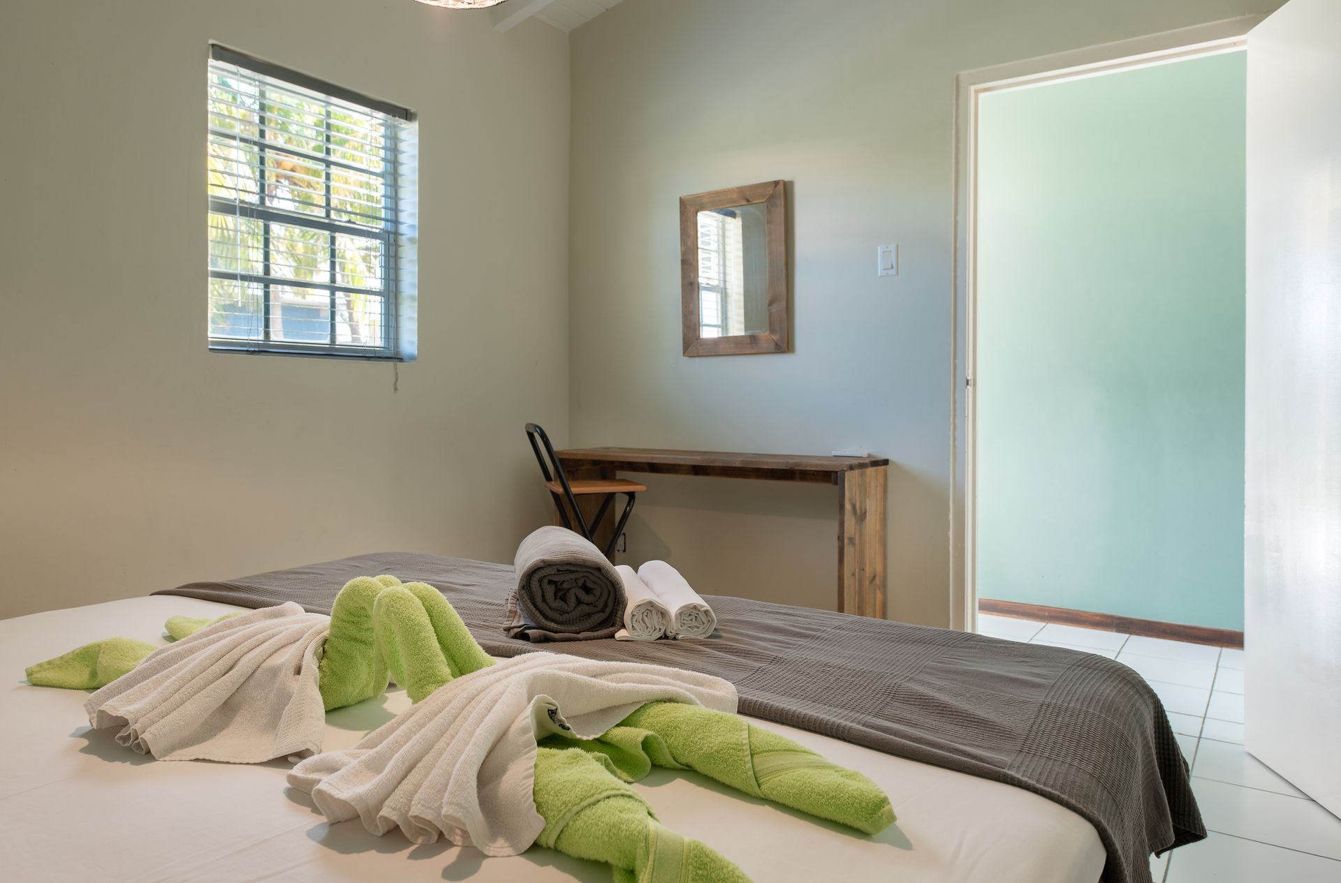 Bedroom with bed, towels, window, desk, and open door. Green and white towels on the bed.