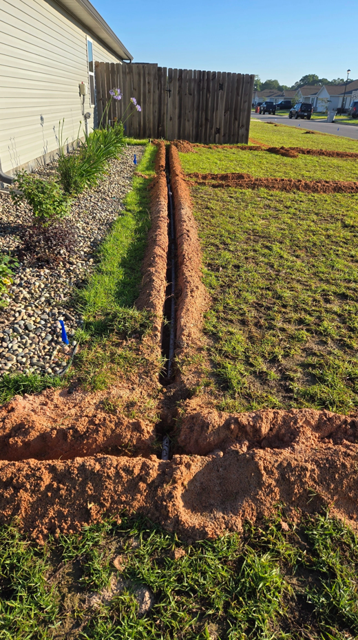 Freshly dug trench in a grassy yard beside a house, with red soil and a wooden fence at the end.