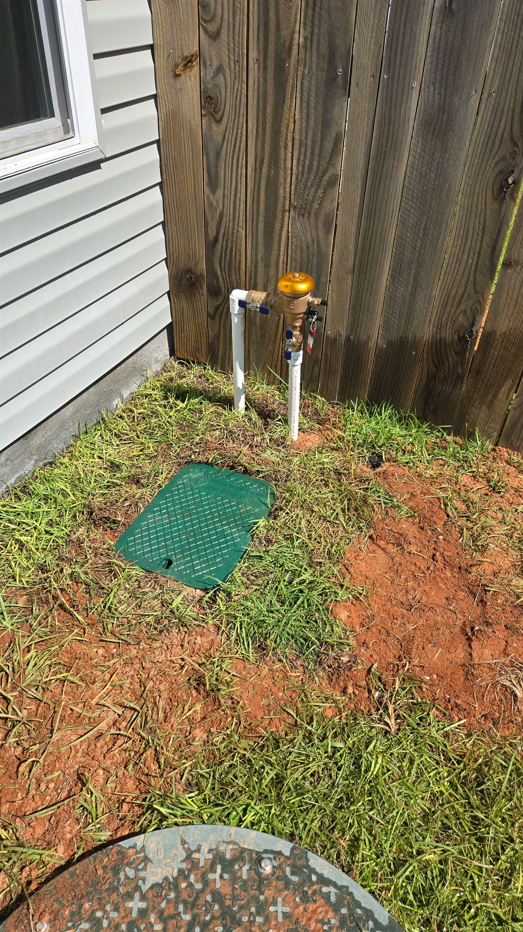 Outdoor utility meters beside a house and wooden fence, with a green utility cover in a patchy yard
