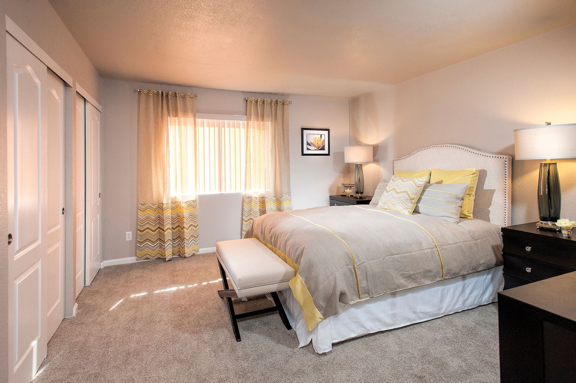 Bedroom with a large bed, pair of nightstands, bench at the foot, and window with patterned curtains.