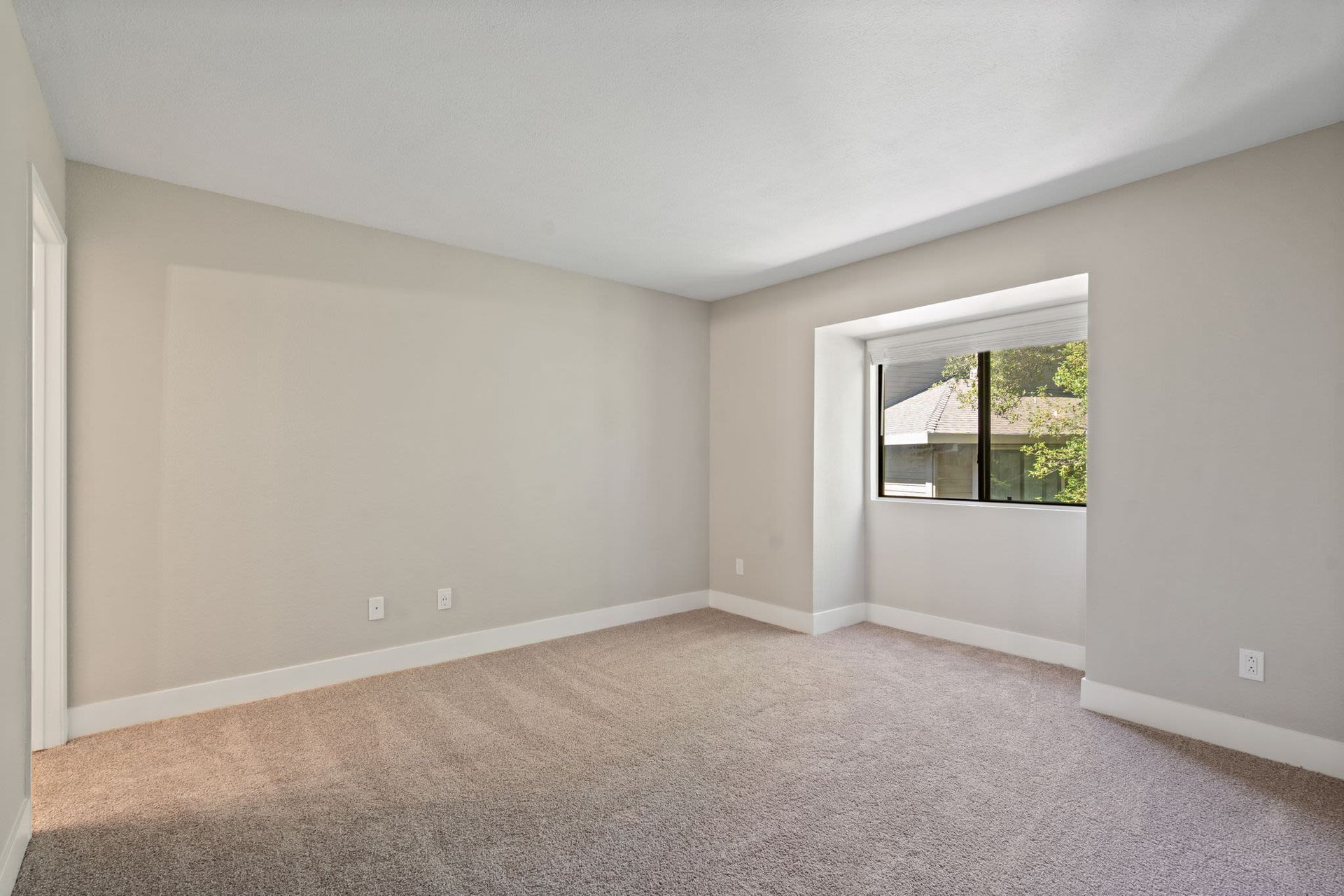 Empty beige-carpeted room with neutral walls and a large window.