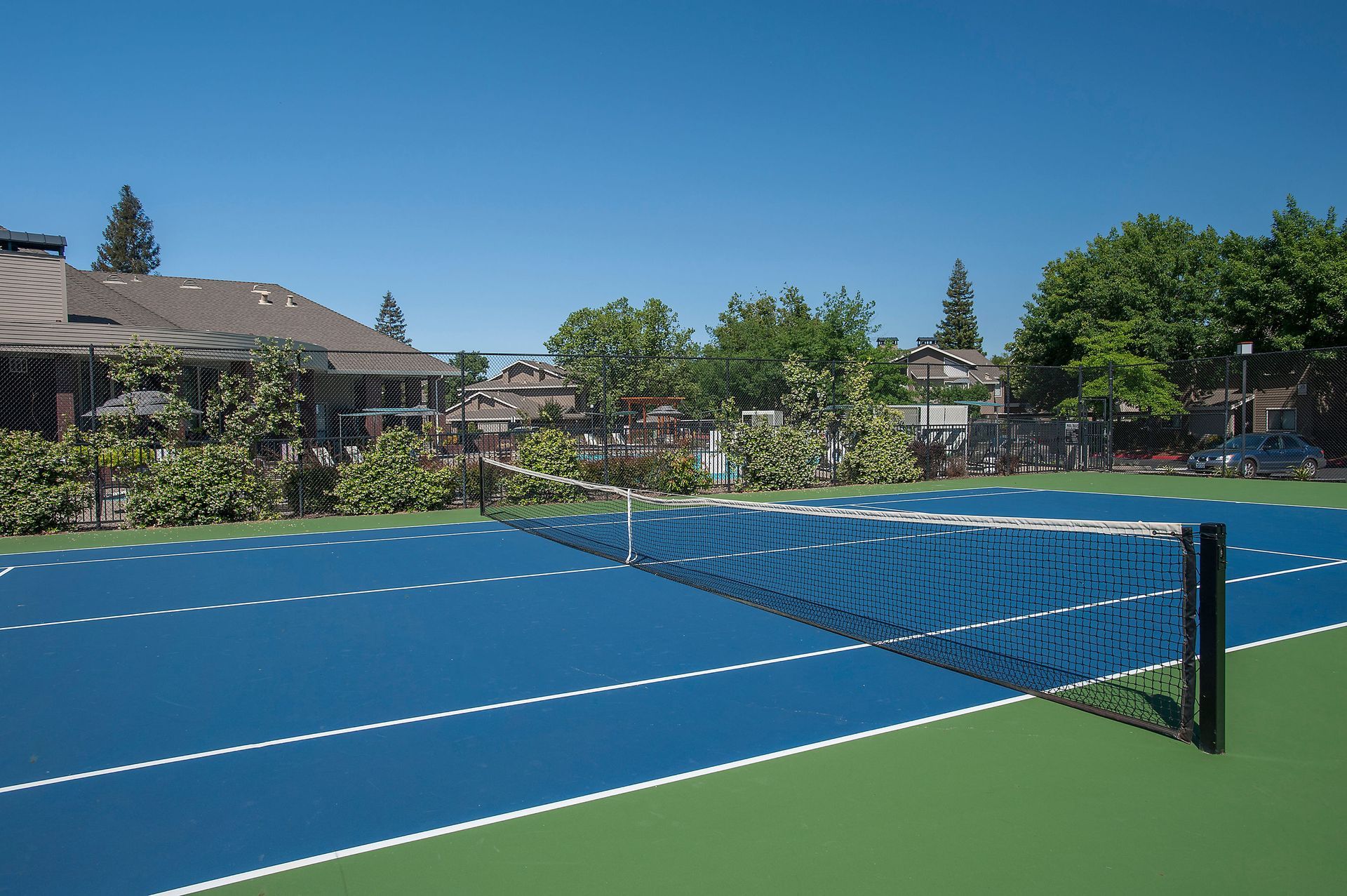 Blue tennis court with net, surrounded by fence and greenery at a residential complex.