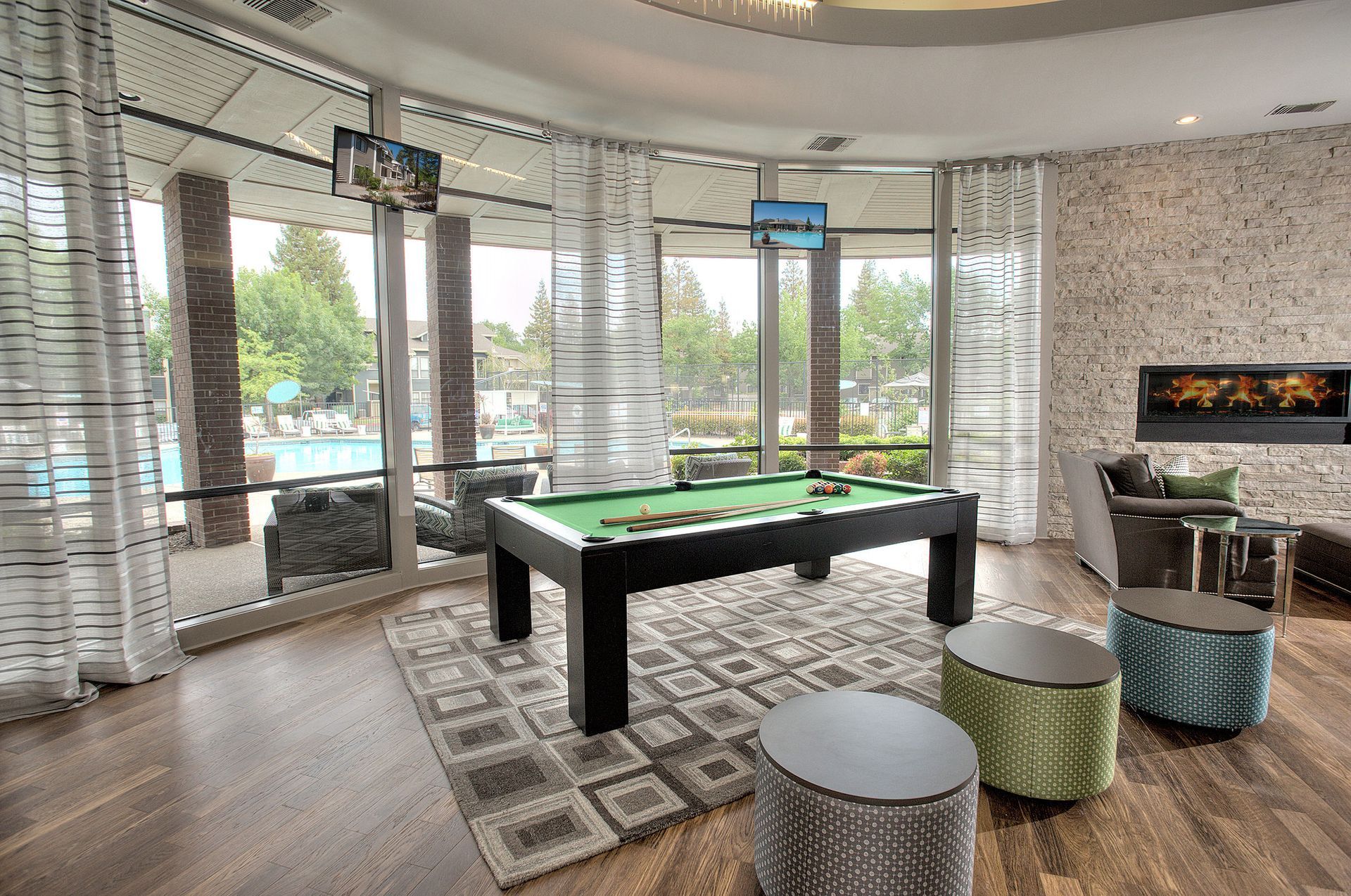 Indoor community lounge with pool table, fireplace, and seating by large windows overlooking the outdoor pool.