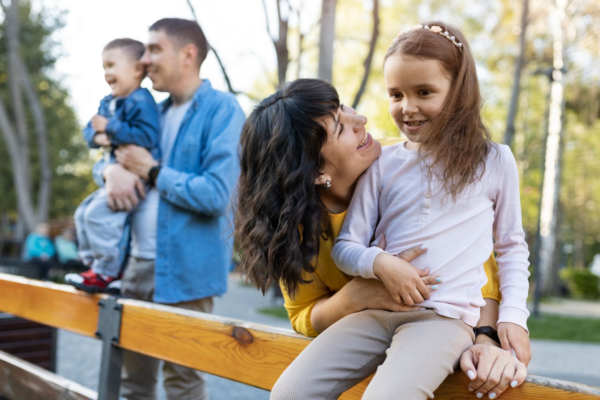 Woman sitting on a bench holding a smiling child, with a man holding a baby in the background.