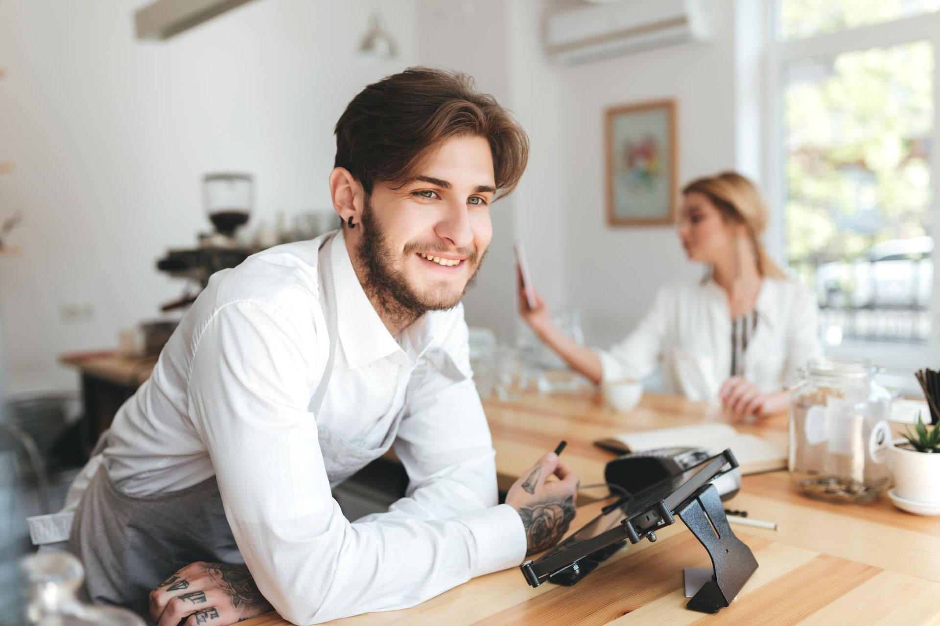 Man smiling at a laptop in a bright café while a woman sits blurred in the background.