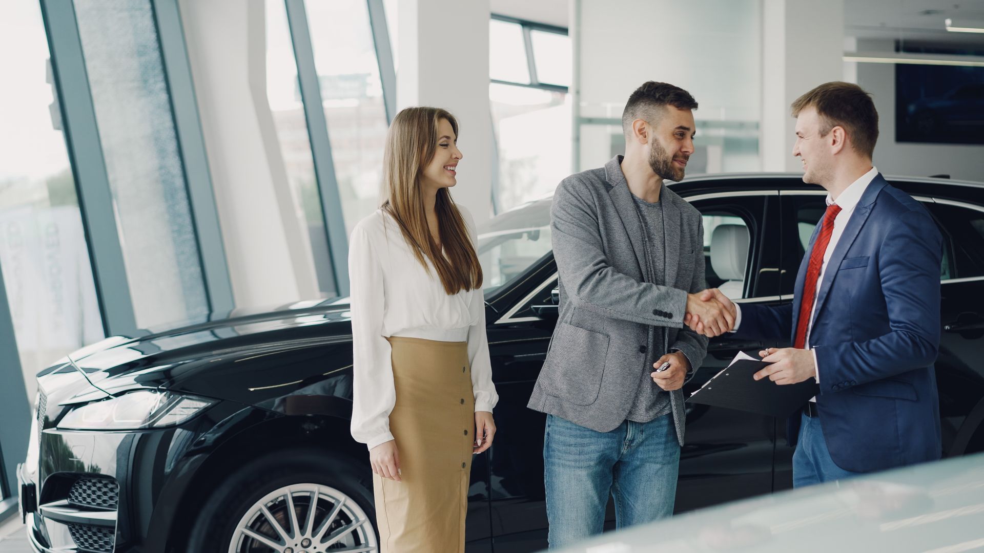 Three people shaking hands beside a black car in a showroom