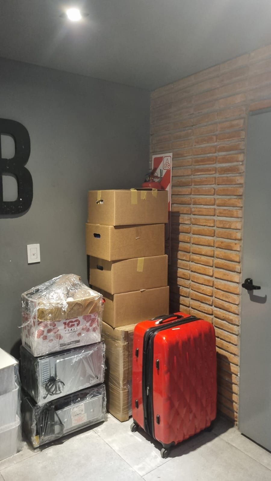 Cajas, una maleta y electrodomésticos apilados contra una pared de ladrillos en un rincón de una habitación.
