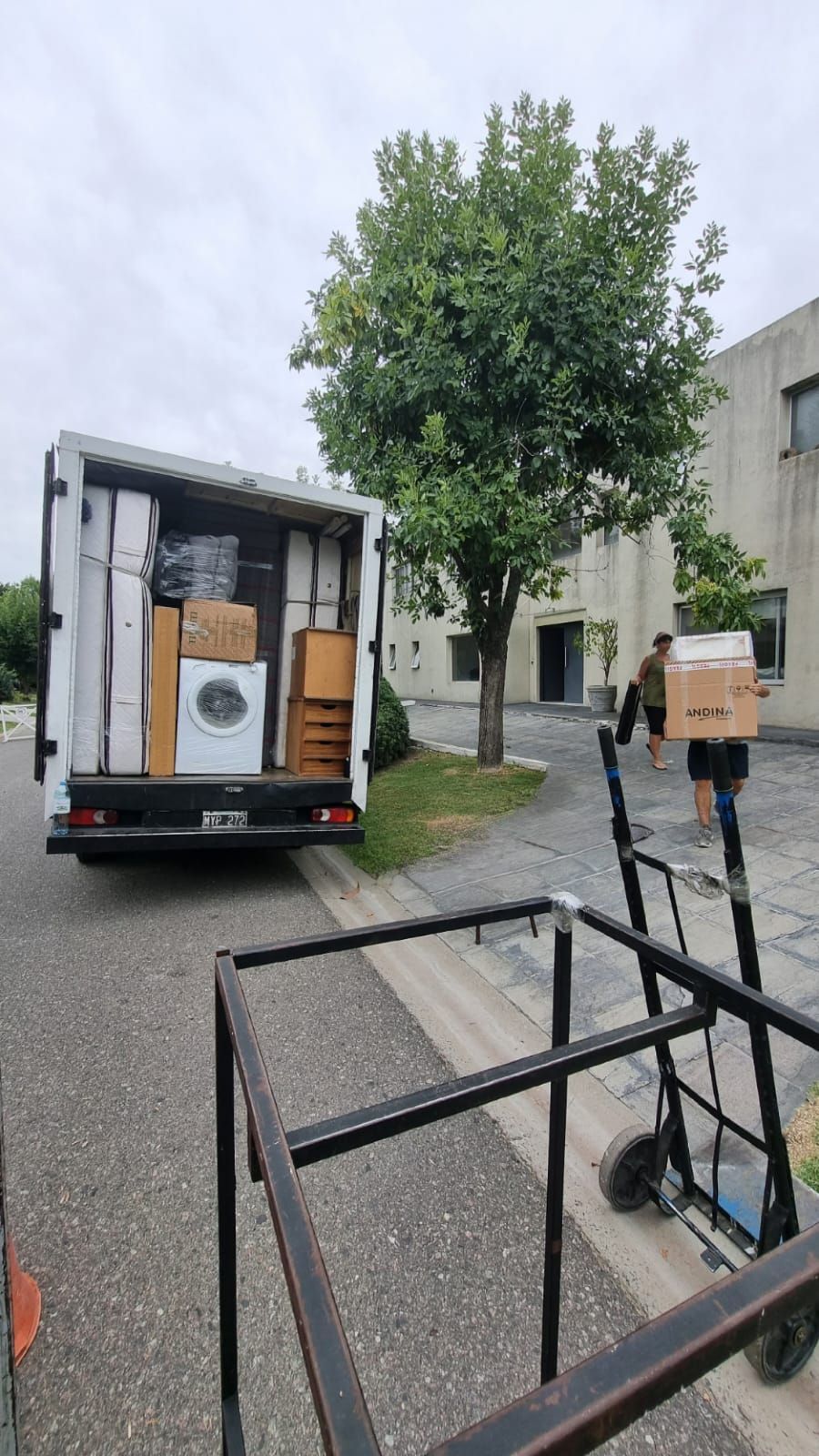 Camión de mudanzas descargando muebles y cajas cerca de la entrada de un edificio. Dos personas cargan cajas.