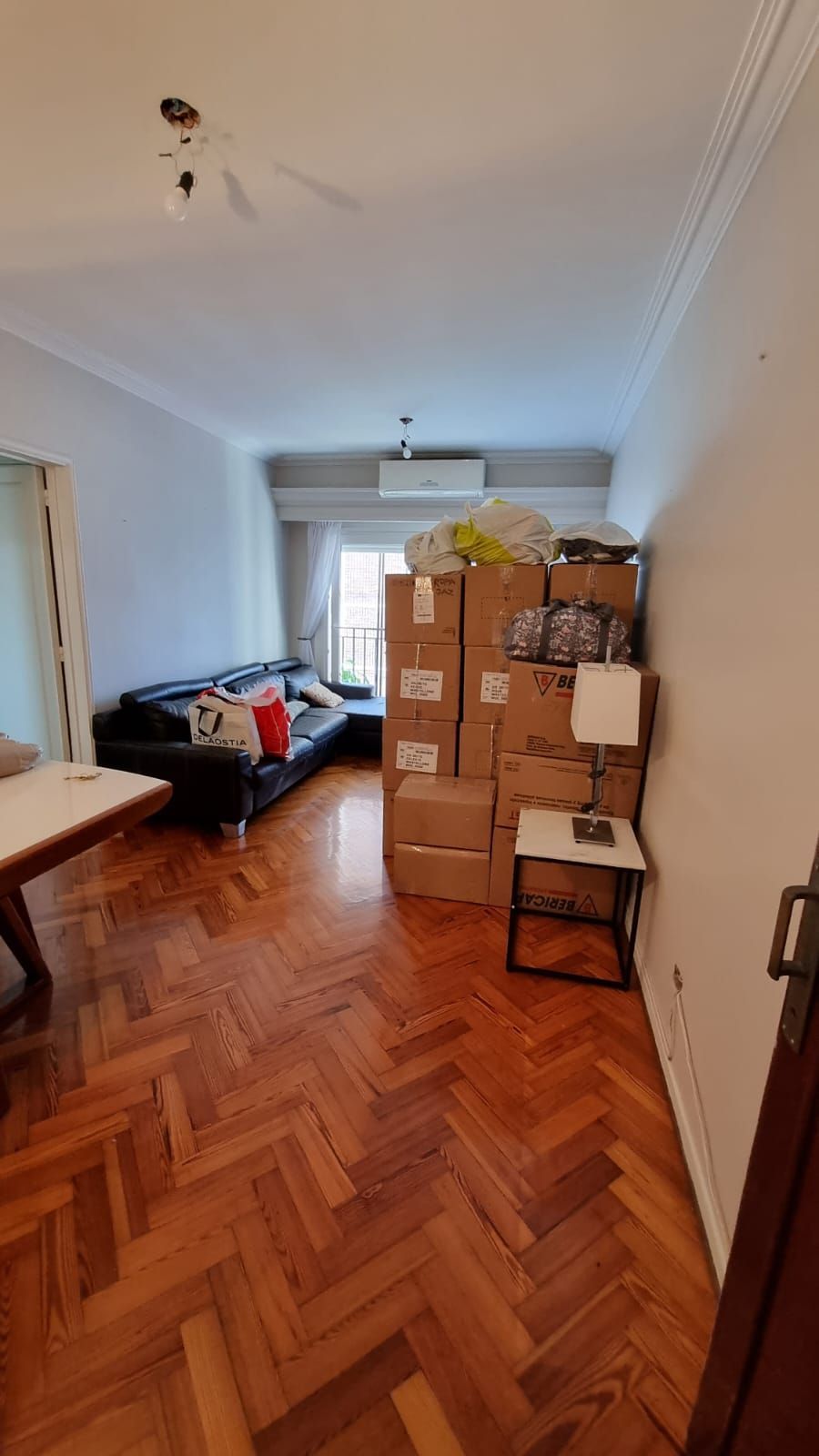 Sala de estar con pisos de madera, sofá negro, cajas apiladas contra una pared y una mesa pequeña.
