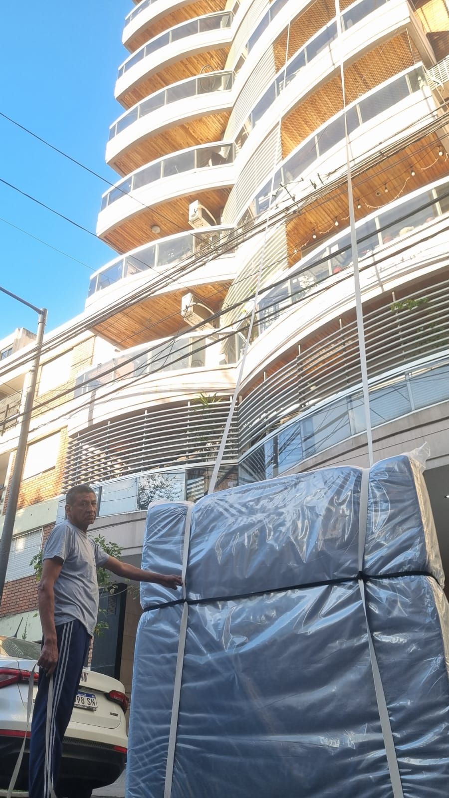 Hombre de pie junto a colchones envueltos frente a un alto edificio de apartamentos.