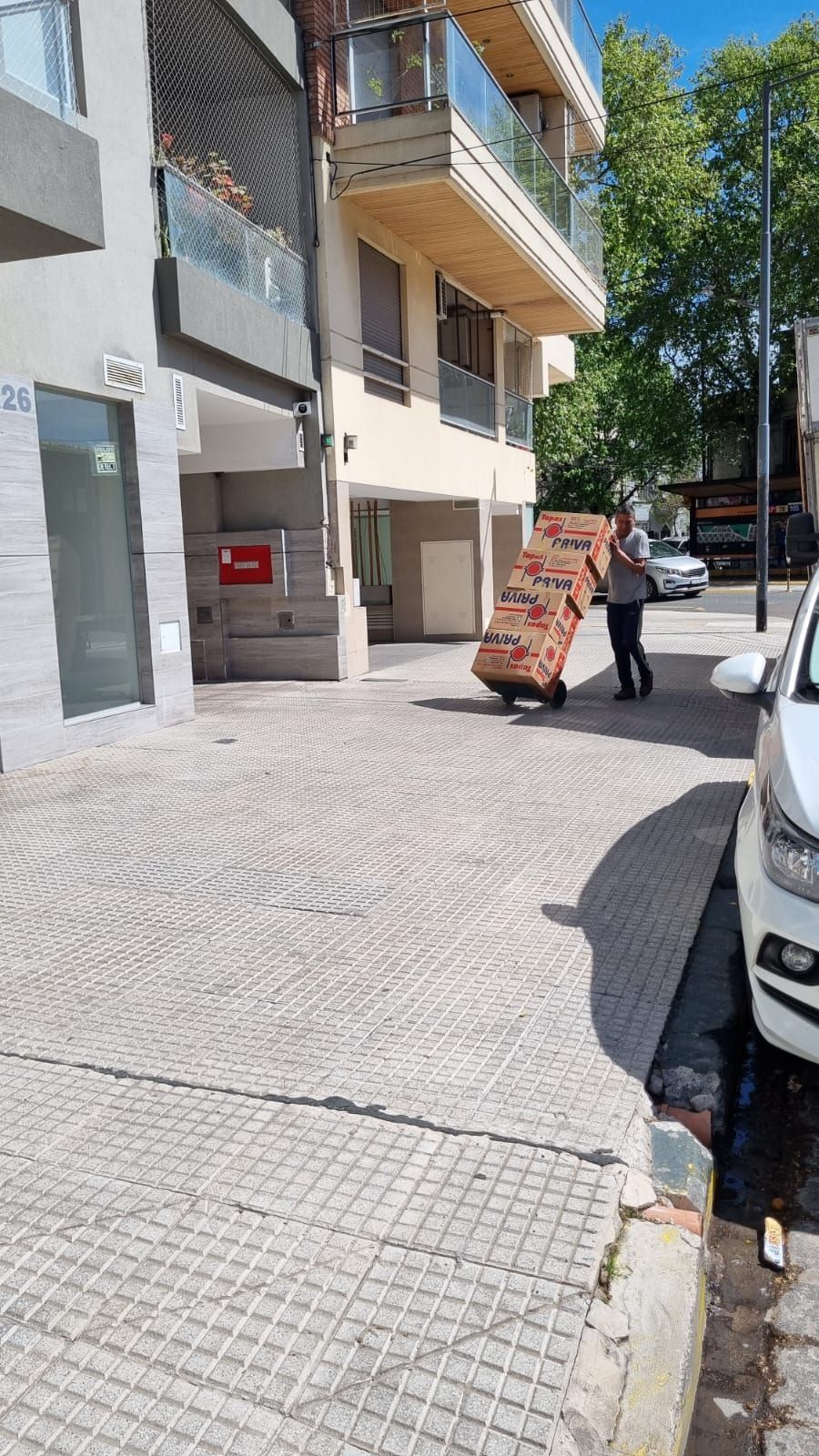 Persona arrastrando un carrito con cajas por una acera de ladrillo. Edificio y coche al fondo.