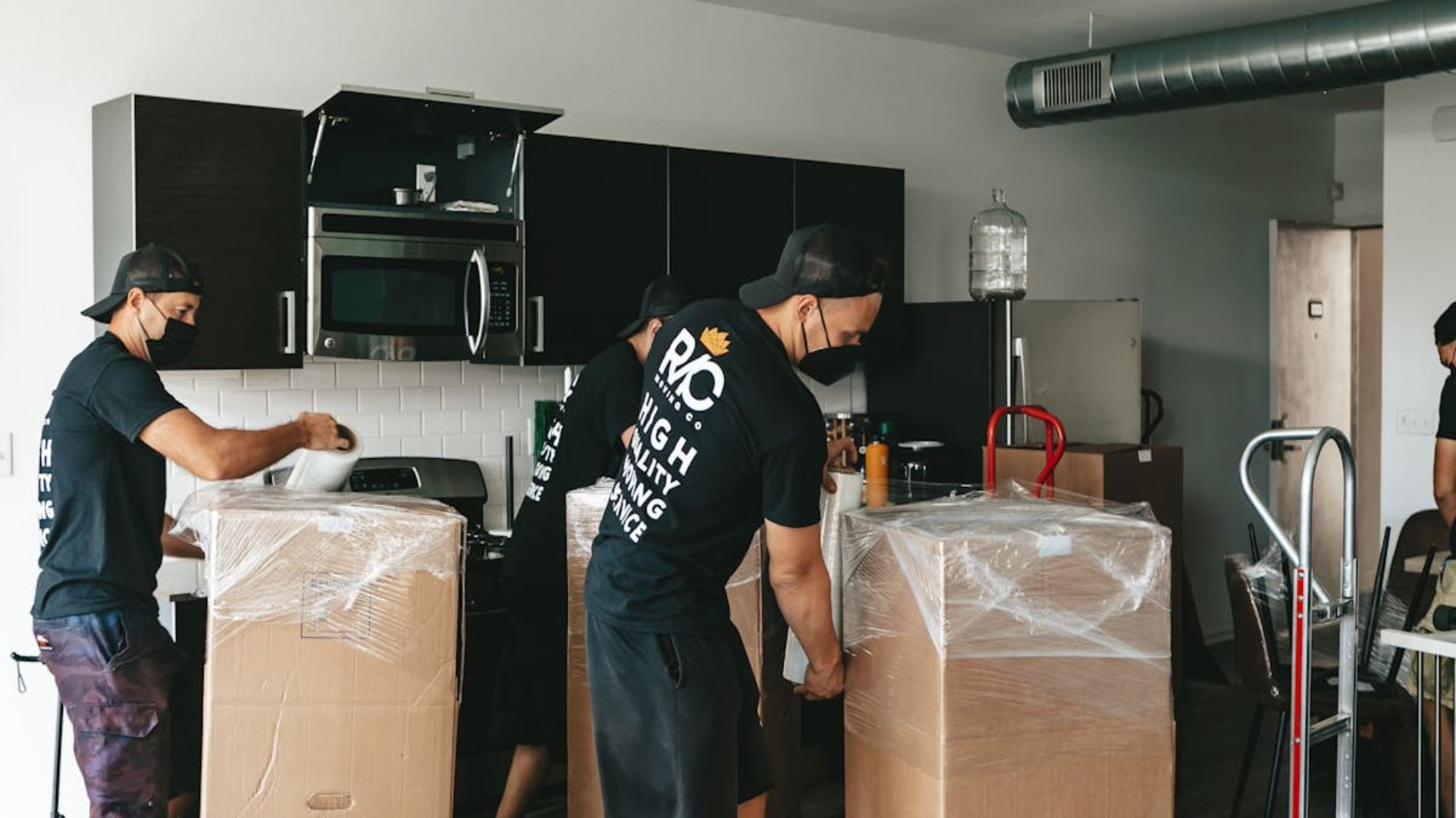 Mudanzas empacando cajas en una cocina. Hombres con mascarillas y camisetas negras, algunas con logotipos, trabajan.