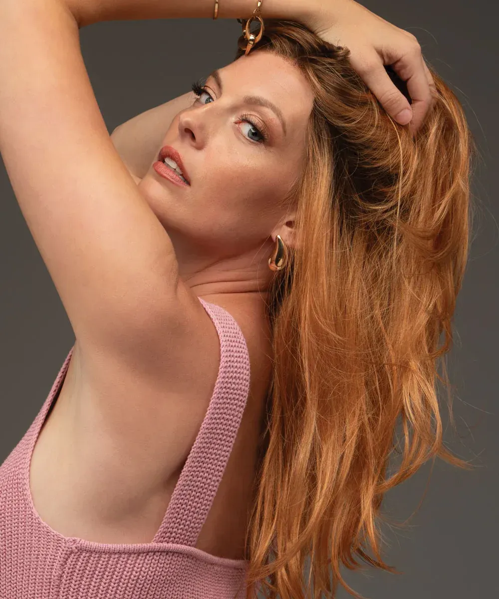 A woman with long red hair is wearing a pink tank top.