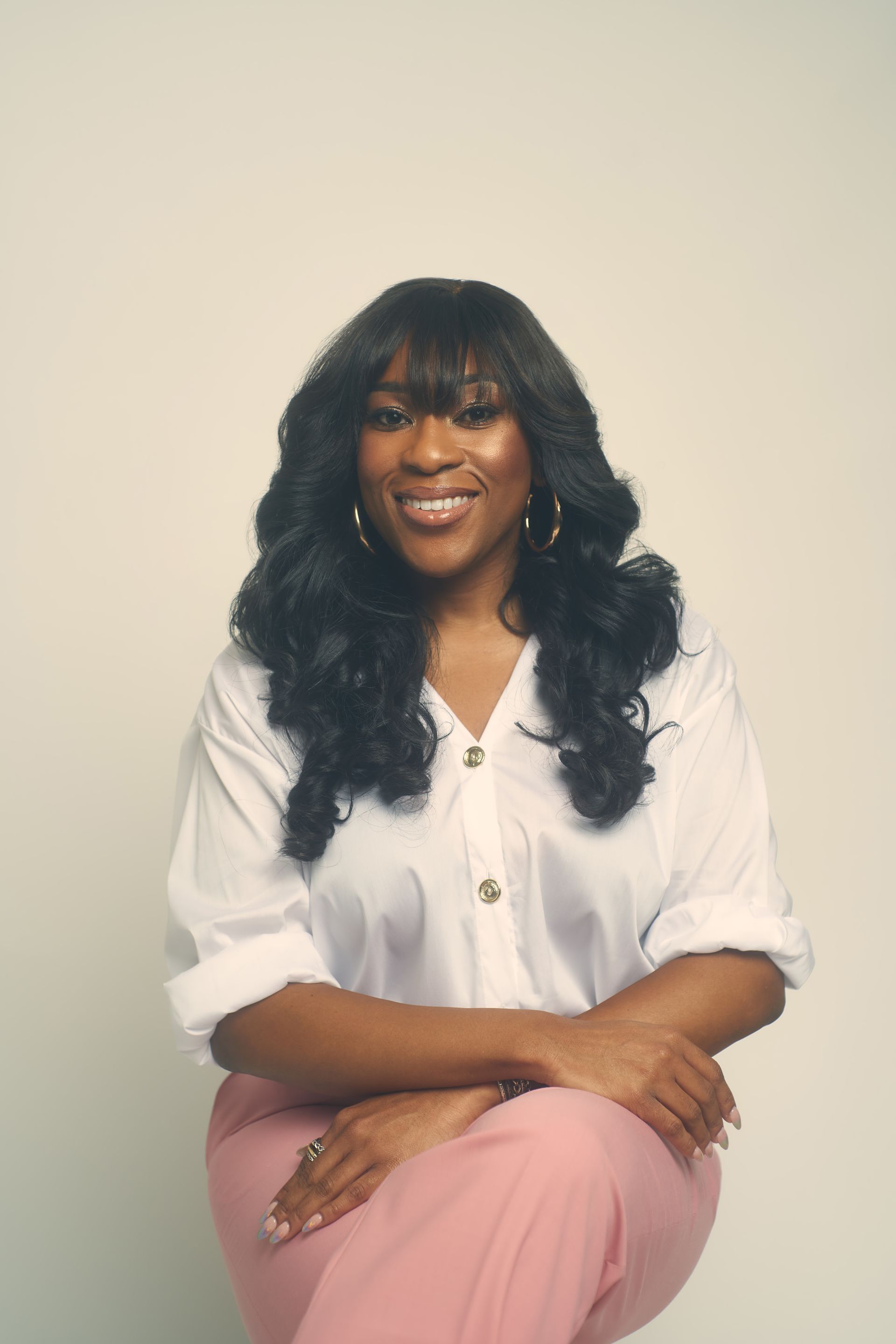Woman with long black hair, wearing white shirt, pink pants, smiling, in front of a white backdrop.
