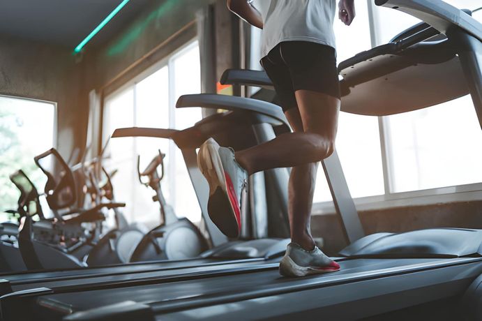 A person runs on a treadmill in a sunlit gym, wearing a white t-shirt and dark shorts.
