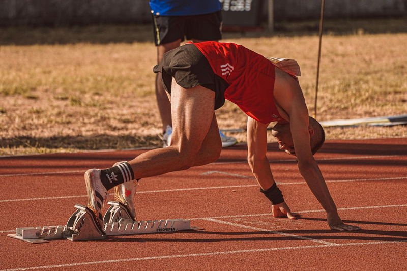 A runner in a red tank top and black shorts in a crouched starting position on a red track with starting blocks.