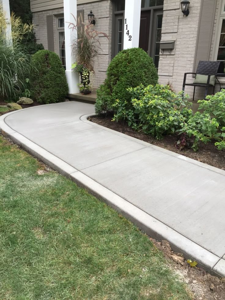 Concrete walkway curving towards a house, lined with green bushes and grass.