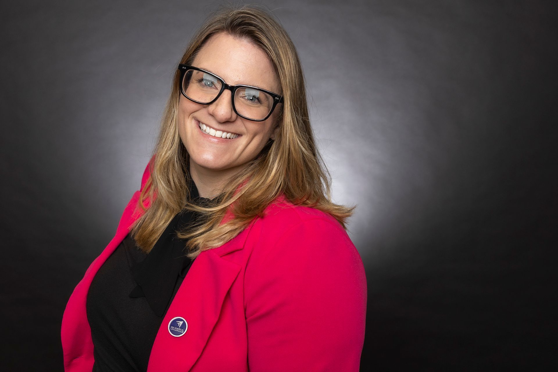 A smiling professional wearing a bright pink blazer and glasses against a dark grey background.